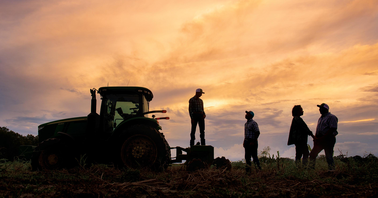 four people stand in silhouette against a sunset with a tractor.