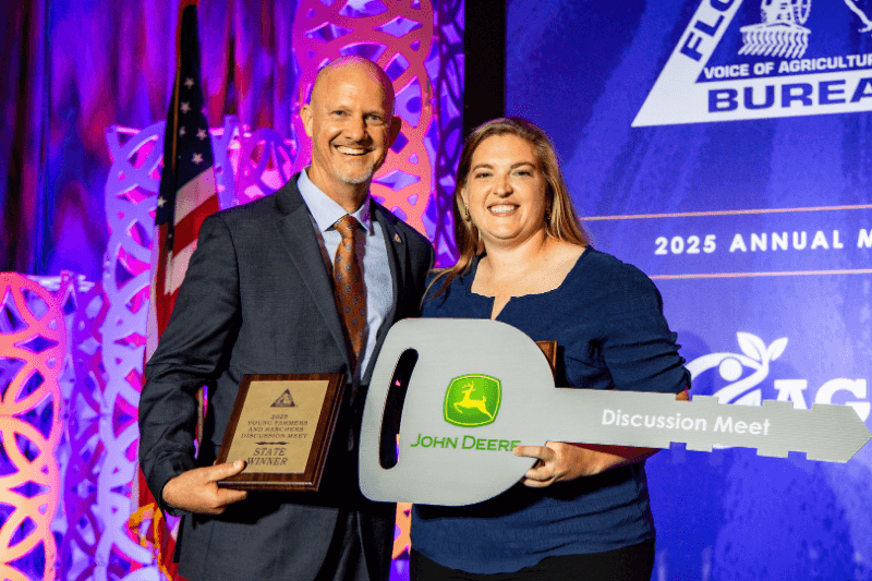 Two people holding an award plaque and a large John Deere Discussion Meet key on a stage