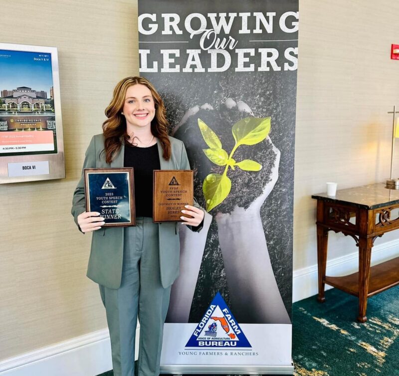 Person holding two Youth Speech Contest award plaques while standing in front of a Florida Farm Bureau Young Farmers and Ranchers banner