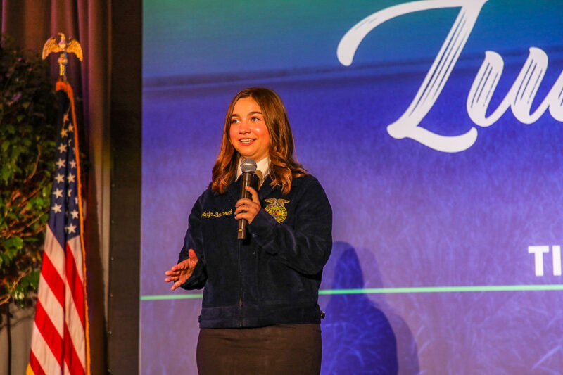 Student speaking into a microphone on stage while wearing an FFA jacket