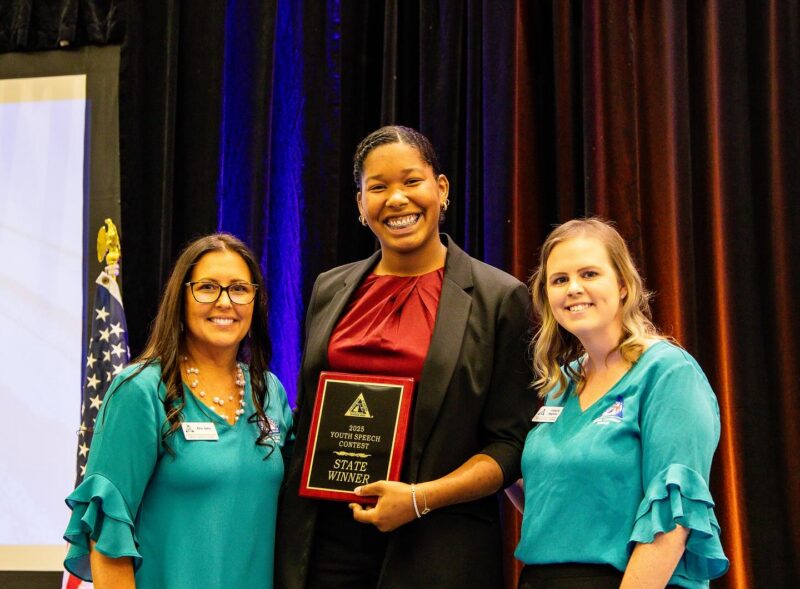 Student holding a Youth Speech Contest state winner plaque while standing with two event representatives on stage