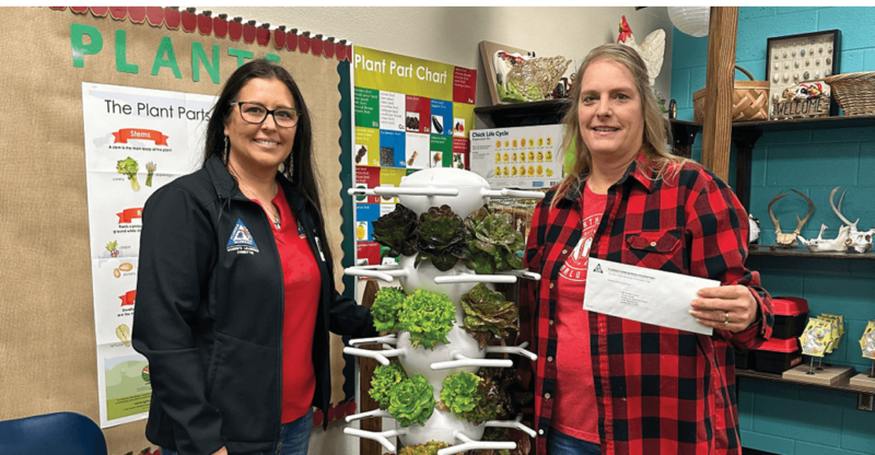 Two educators stand in front of a hydroponic garden setup in a classroom.