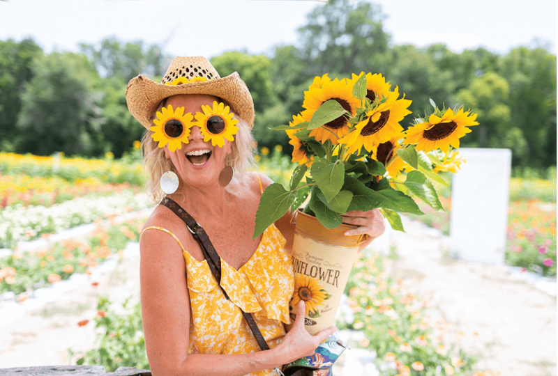 A caucasian person in a cowboy hat holds a bouquet of sunflowers. They are smiling at the camera and have matching sunflower sunglasses.