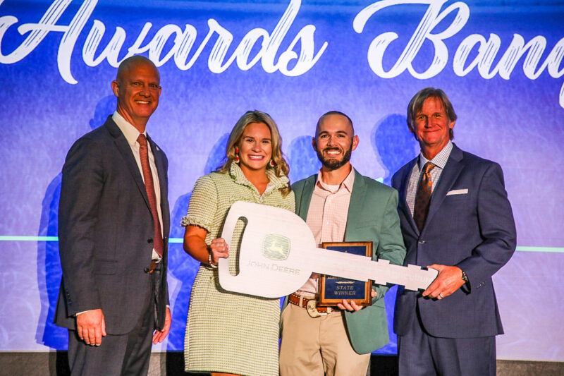 Four people standing on a stage holding an award plaque and a large John Deere key during an awards presentation