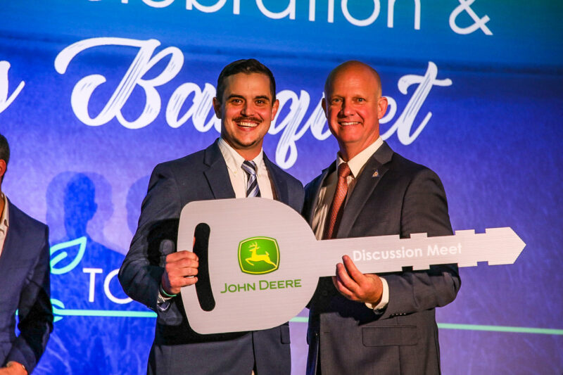 Two people standing on a stage holding a large John Deere key labeled “Discussion Meet”