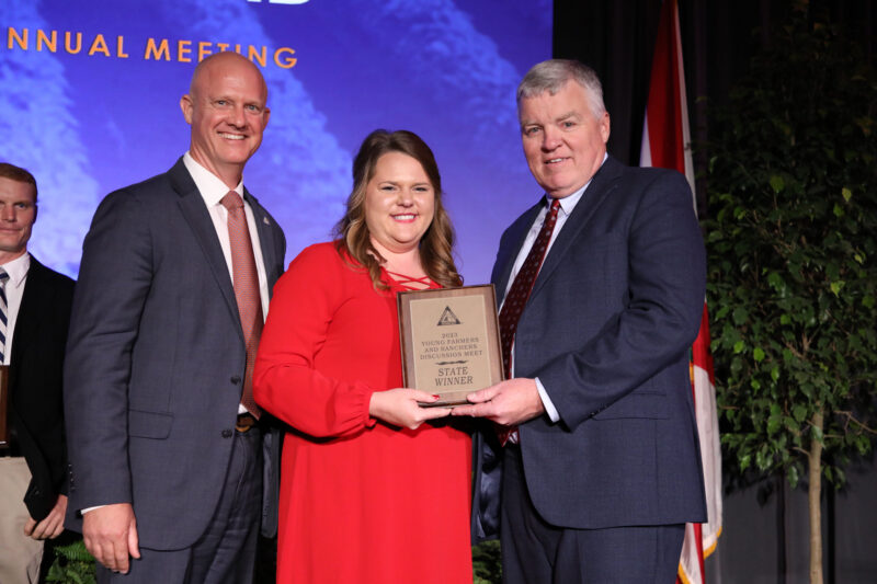 Three people standing on a stage holding a state winner award plaque during a Florida Farm Bureau event