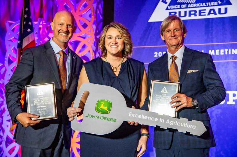 Three people standing on a stage holding award plaques and a large key labeled “John Deere Excellence in Agriculture”