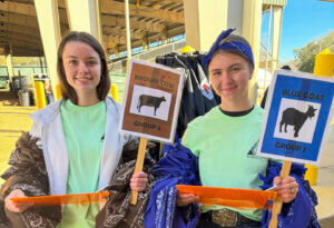 Two participants hold group signs labeled “Brown Cow Group 3” and “Blue Goat Group 7” during an agricultural education activity in a covered barn.
