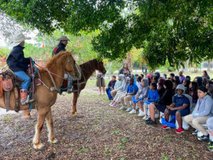 Two people on horseback speak to a seated group of students during an outdoor educational demonstration under a large tree.