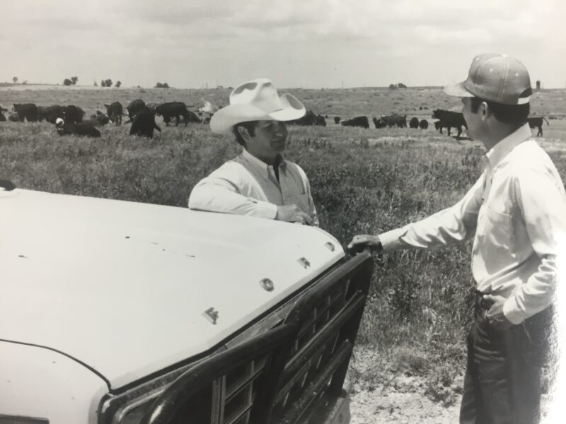 Two people leaning against a pickup truck while talking in a pasture with cattle in the background