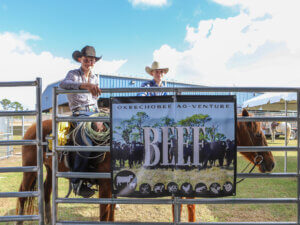 Two participants seated on horses behind a metal fence hold a banner reading “Okeechobee Ag‑Venture BEEF” at an agricultural education event.