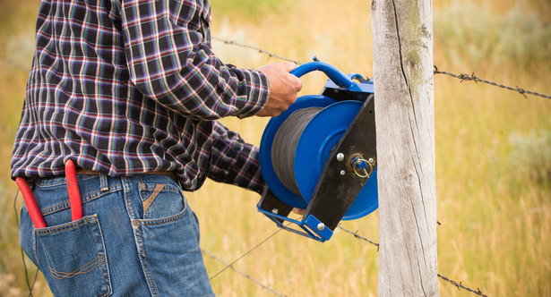 Person installing barbed wire fencing using a blue wire reel mounted on a fence post in a rural field.