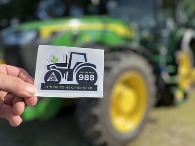 Hand holding a sticker with a tractor icon and the message “Call or text 988 – It’s OK to ask for help,” with a blurred green tractor in the background.